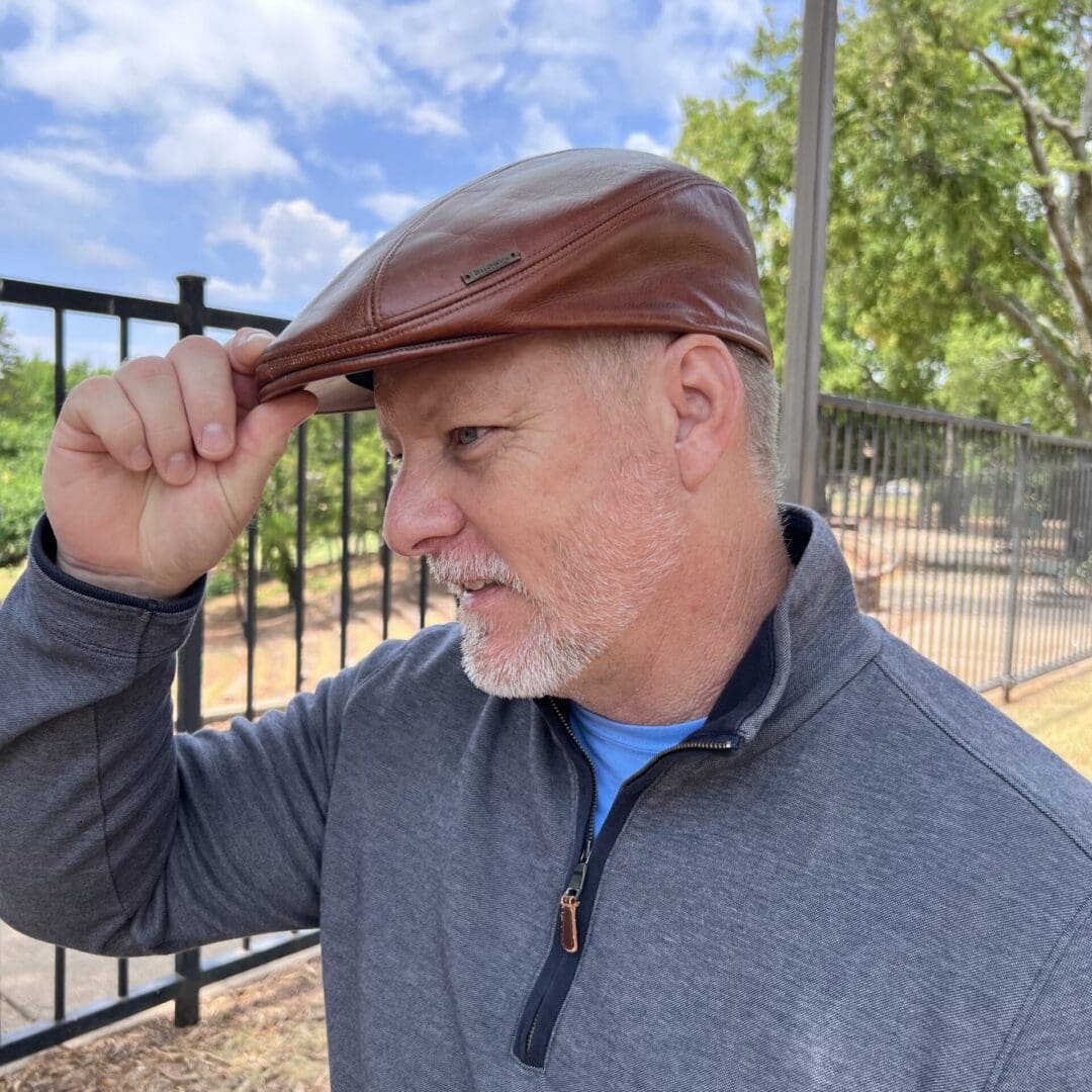 A man in grey jacket holding his hat up to the side of his head.